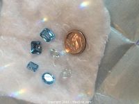 Photo showing six loose blue topaz gemstones with varying cuts and sizes along with two smaller clear gemstones on white background next to a dime for size reference.
