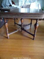 Front view of hardwood dining table showing turned legs, cross supports, and two connected leaves. Visible wear and scratches on surface, legs show some signs of age.