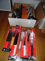 Photo showing knives in red packaging with white handles and a meat cleaver with a yellow handle on a floor in front of a drawer with more utensils inside.