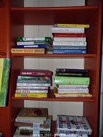 Wide shot of multiple shelves filled with assorted cookbooks, showing stack arrangement and identifiable titles revealing collection size and themes.