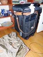 Photo of black Eddie Bauer rolling suitcase with brown leather strap, olive garment bag on floor, and wooden shelving in background.