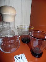 Photo showing clear Danish glass decanter with rounded shape and natural wood stopper, alongside two art glass drinking glasses with tinted rims and black bases.