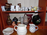 Full view of two wooden shelving units filled with over 60 ceramic and pottery items including pitchers, jugs, baking ramekins, egg cups, and vases in a mixture of colors including white, brown, rustic earth tones, and floral patterns.