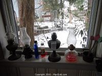 Four vintage oil lamps of various sizes and two small bottles on windowsill with snow visible outside. Black metal bases and glass chimneys visible.