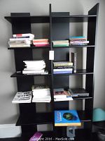 Front view of one black pressed wood bookcase piece with books on shelves showing six shelves, vertical dividers, and open back design.