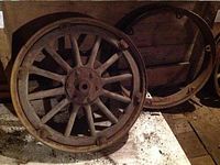 View showing a wooden spoke wheel with a detached metal rim leaning against a wall on a white surface, highlighting the condition and detailing of the wheel and rim.