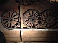 Three wooden spoked wheels arranged on a wooden rack in a dimly lit barn or shed, showing overall condition including surface wear and aging