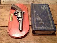 Photo of decorative plaque displaying a metal replica of a Smith & Wesson Model 36 revolver mounted on reddish wood base next to an empty Smith & Wesson branded blue box.