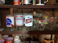 Front view of 10 vintage beer glasses on wooden shelf showing Pabst Blue Ribbon, Budweiser, Schlitz logos and other beer brandings.