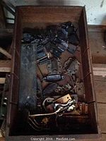 Wooden box filled with assorted door hinges, metal mounting plates, black metal plates, and various door handle parts and coat hooks, showing mixed metal finishes and patina.