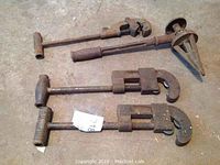 Four antique pipe cutting tools arranged on ground showing wooden and metal parts with rust and patina.
