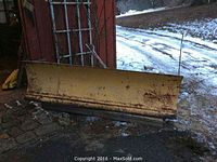 Full view of yellow snow plow blade leaning against a structure outside on a paved surface with some snow and ice nearby. Shows the size and form of the blade.