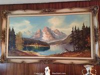 Framed landscape painting showing the overall artwork and frame against a wood-paneled wall, highlighting the decorative frame and mountain lake scene.