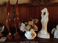 Wooden crane bird sculptures, porcelain floral vase with faux roses, white ceramic statue of an embracing couple, and cherub figurine on wooden shelf