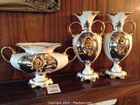 Three matching Italian porcelain vases arranged on a wooden surface, two tall and one short, with gold accents and floral appliques.