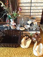 Wide view of assorted glass vases, candle holders, ceramic swans, wood vase, and wicker basket arranged on a dark surface with a fireplace background.
