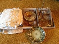 Photo showing assortment of linens folded on left and various glass and metal decorative items in boxes on the right, including candle holders and amber glass pieces.