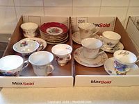 Photo showing two boxes containing 12 assorted tea cups and saucers with floral and decorative patterns in various colors and styles.