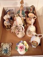 Assorted decorative porcelain figurines and ceramic pieces including girls, pitchers, floral vases and a framed religious picture, all arranged in a cardboard box.