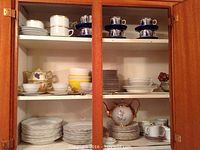 Open wooden cabinet showing multiple shelves of assorted plates, bowls, tea cups, saucers, and a decorative teapot. Porcelain and china in various patterns including floral and traditional designs.