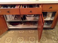 Kitchen cabinet with drawers open showing various kitchenware items inside including cutlery in upper drawers, mixing bowls, and CorningWare dishes on shelves.