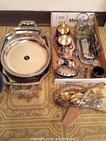 Wide shot of assorted silver plate serving trays, salt and pepper caddy, utensils, and accessories arranged in a box and on a small pedestal