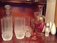 Photo showing two decanters, one clear cut glass and one pink with gold decoration, along with matching clear cut glass highball or water glasses, and small amber-colored glasses.