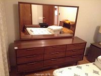 Full view of walnut-tone wood dresser with nine drawers topped by matching rectangular framed mirror in bedroom setting