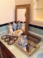 Overall view of all items on bathroom countertop including ornate mirror, porcelain figurines, decorative shell bowl, perfume tray, and floral decor pieces