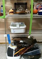 Metal grill pans and skewers with wooden handles piled on a metal shelf along with a plastic container and other kitchen items including glass containers.