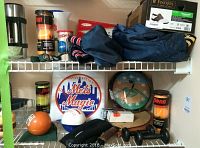 Mets baseball themed items on wire shelf including two outdoor seat cushions, baseball clock in green frame, two clear plastic pitchers, bank cap, two black hand weights and Pinnacle golf balls box.