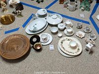 Array of assorted porcelain and china cups, saucers, platters and child dinner set, arranged on floor within blue tape boundaries, showing variety of items in lot.
