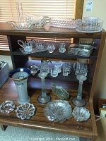 Wide shot of multiple crystal and glass pieces arranged on a wooden shelf including bowls, candlesticks, vase, creamer, and other serving pieces.