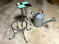 Photo showing two iron garden stands and a galvanized watering can on concrete floor