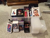 Plastic containers filled with a mix of VHS tapes and DVDs, arranged with visible titles including Bob Dylan, Metallica, and movies.