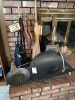 Photo showing fireplace tools on stand, a broom, wooden bellows, metal firewood basket, and a metal scoop on a brick hearth in front of a glass fireplace enclosure.