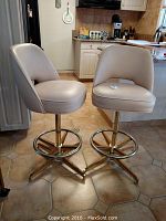 Pair of two vinyl upholstered stools with light beige cushioned seats and backs, metal chrome bases with footrests, shown in kitchen setting