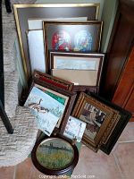 Photo showing a group of various framed prints and paintings stacked against a wall, including assorted sizes and frame styles.