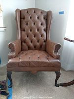 Front view of the wingback chair showing brown vinyl upholstery, deep button tufting, rolled arms with nailhead trim, and wooden cabriole legs.
