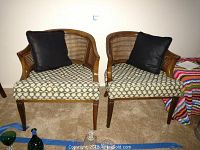 Two vintage wooden armchairs with caned backs and light upholstery featuring a leaf pattern, each with a black throw pillow on the seat.