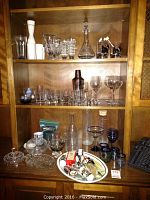 Photo of wooden cabinet shelves with glass decanters, stemware, cocktail shaker, blue glasses, and white sake set.