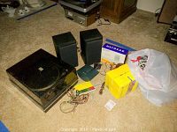 Wide angle photo showing turntable, speakers, boxes, cables, and a plastic bag with extra items on carpeted floor.
