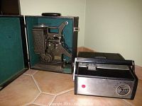 Keystone M-8 8mm film projector in open carrying case alongside Bell & Howell Telesonic machine on tile floor.