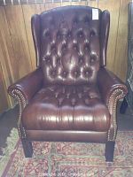 Front view of brown leather wingback chair with tufted back and seat, and studded nailhead trim along arms.