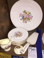 Top-down photo showing cream pitcher, stacked plates with floral designs and gold trim, and serving spatula on wooden surface.