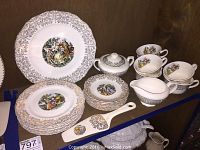 Full view of plates, teacups, cream and sugar bowls, and serving items showing gold decoration and colorful central art.