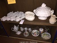 Top shelf showing white ornate tureen, gravy boats and creamer, with bottom shelf showing small teapot, miniature cups, and decorative plates.