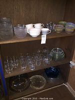 Shelf showing various glass bowls, white ceramic cups, clear glasses, and plates in wood shelving unit