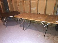 Two folding tables, one circular with dark brown top and metal legs, other rectangular with wood pattern top and metal legs, shown on concrete floor with exposed wall studs behind.