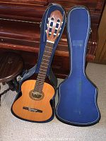 Classical acoustic guitar next to blue soft case on carpeted floor.